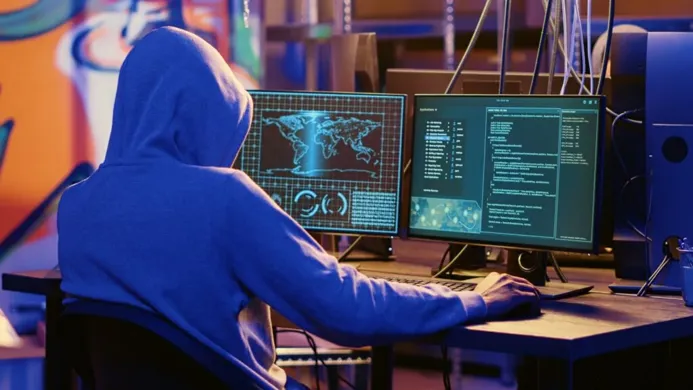A person in a hoodie working on multiple computer screens displaying code and a world map in a dark room with ambient lighting.