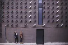Two individuals standing in front of a wall covered with various security cameras, showcasing an urban art installation.