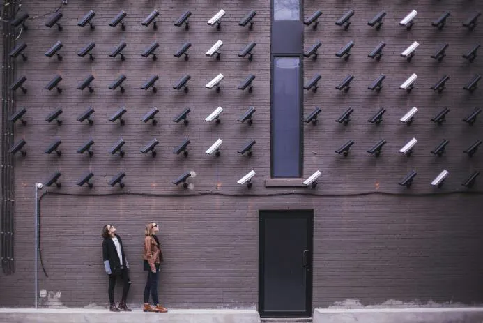 Two individuals standing in front of a wall covered with various security cameras, showcasing an urban art installation.